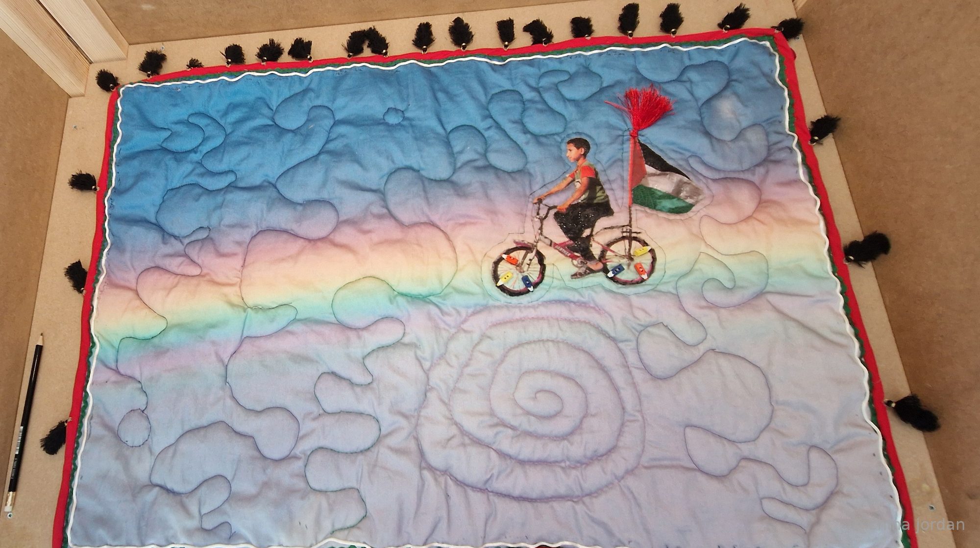 Dream — quilt of a child riding a bicycle with a Palestinian flag across a rainbow sky